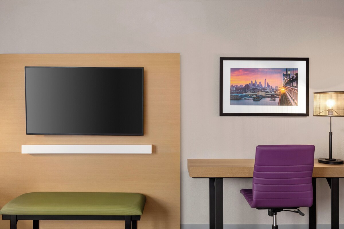 A hotel room with a flat-screen TV mounted on the wall, a framed cityscape picture, a desk with a purple chair, and a green bench.