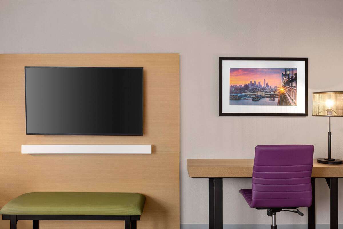 A hotel room with a flat-screen TV mounted on the wall, a framed cityscape picture, a desk with a purple chair, and a green bench.
