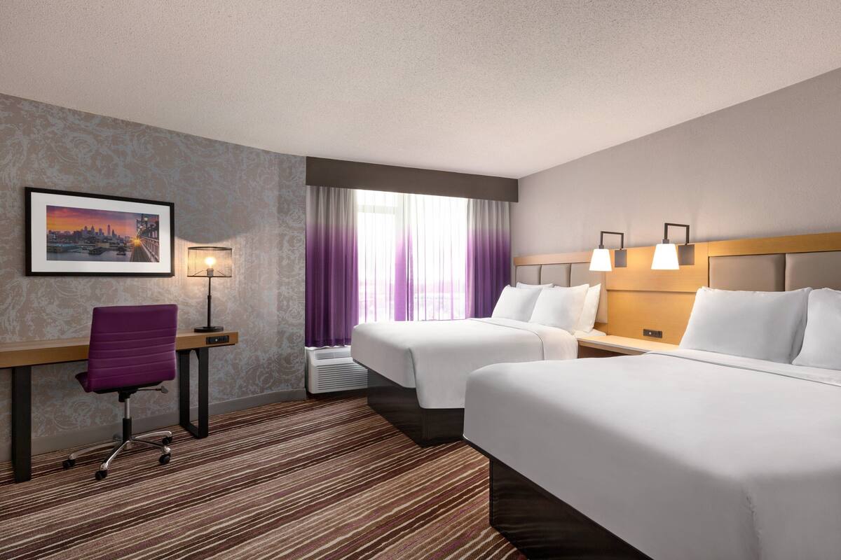 A hotel room with two beds, a desk, a chair, and a large window with purple curtains.