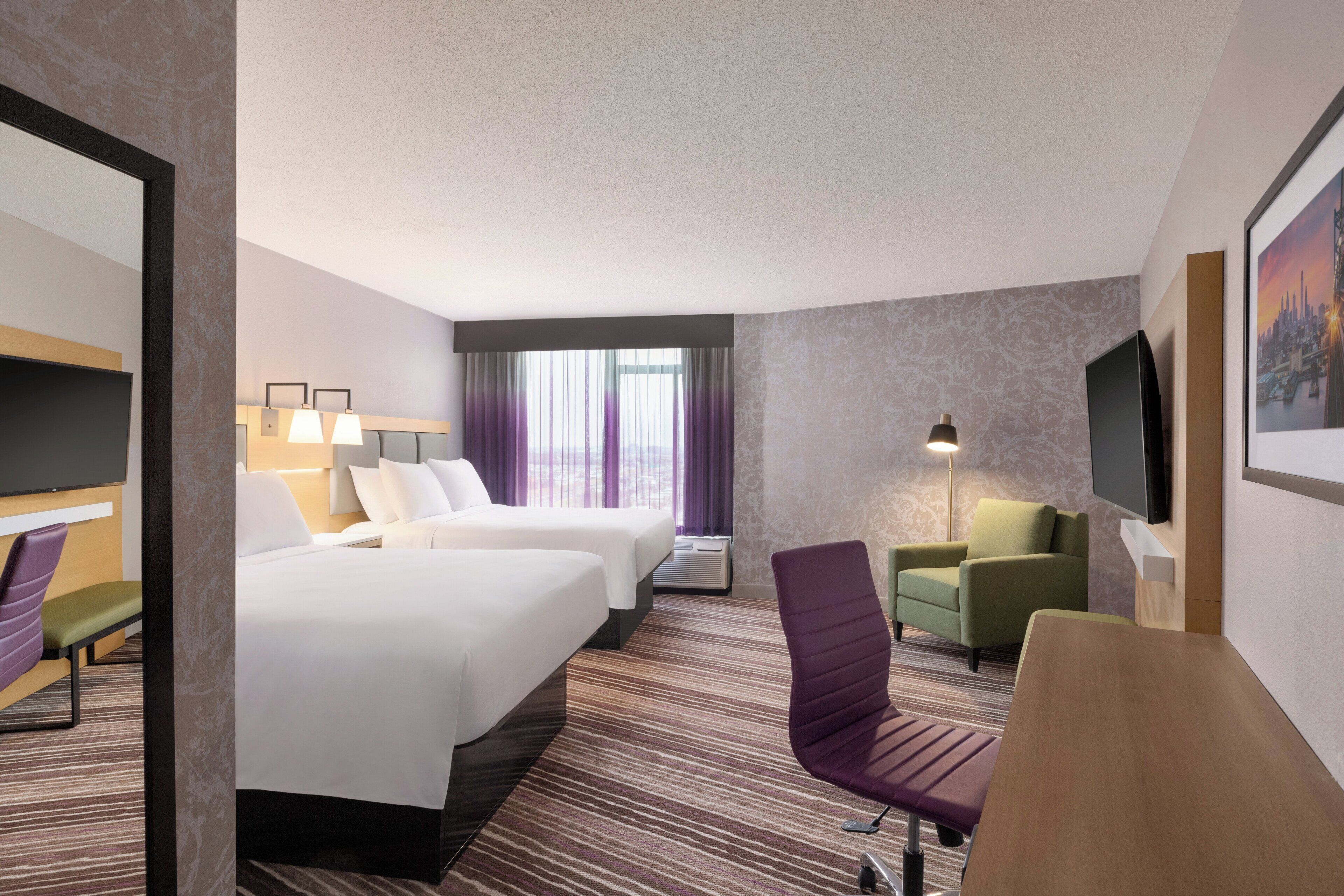 A hotel room with two beds, a desk, a chair, a TV, and a window with curtains.