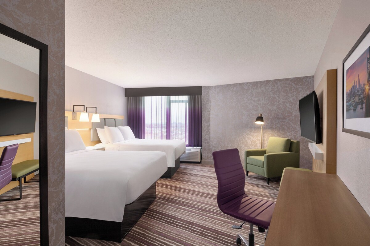 A hotel room with two beds, a desk, a chair, a TV, and a window with curtains.