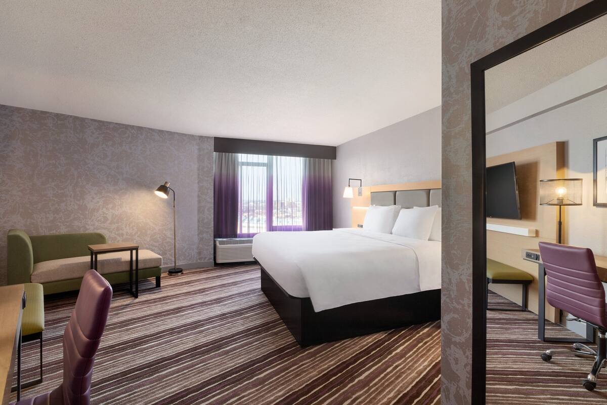A hotel room with a bed, a desk, a chair, a sofa, a lamp, and a large mirror.