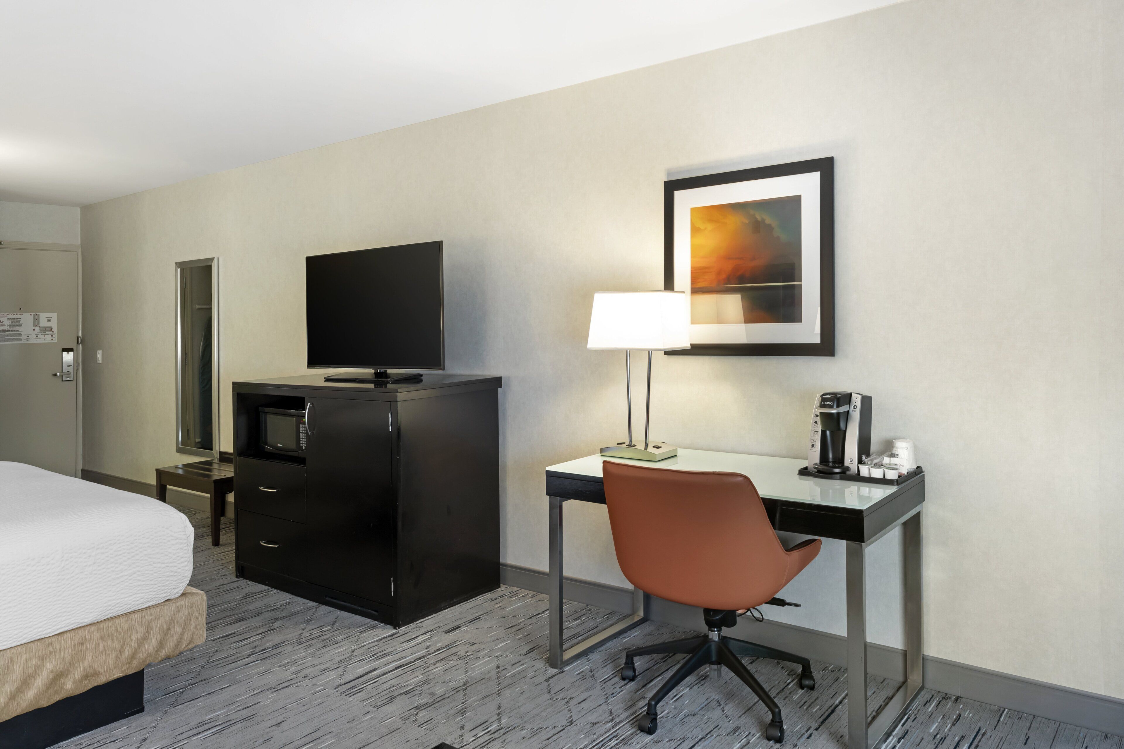 A hotel room with a bed, a desk with a chair, a television, a coffee maker, and a framed picture on the wall.