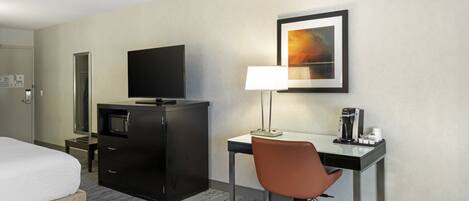 Habitación de hotel con cama, escritorio con silla, televisor, cafetera y un cuadro en la pared.