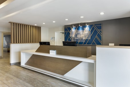 A modern reception desk with a wooden accent, a blue geometric wall design, and a sign indicating the hotel name.