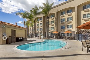 A swimming pool with lounge chairs and umbrellas, surrounded by palm trees and a building with multiple windows.
