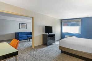 A hotel room with a bed, a desk, an orange chair, a blue sofa, a television, and a window with blinds.