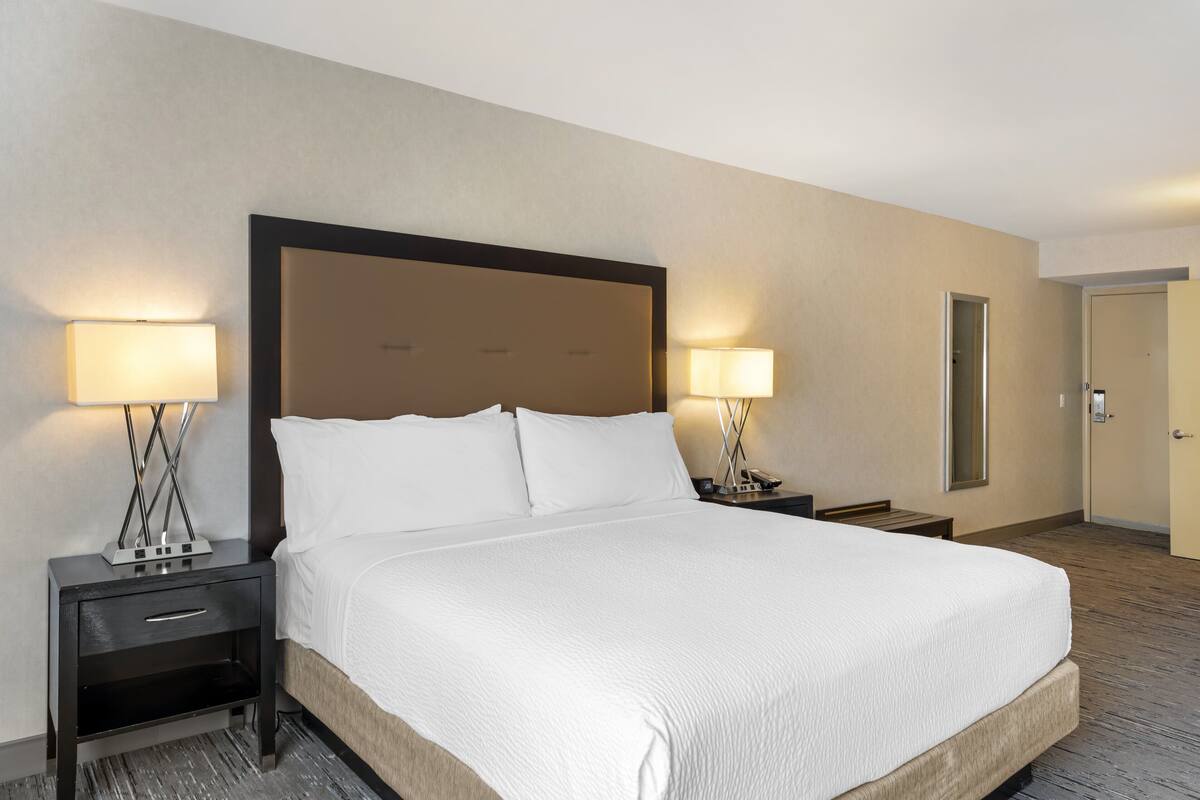 A hotel room with a large bed, two bedside tables with lamps, a nightstand, a mirror, and a door.