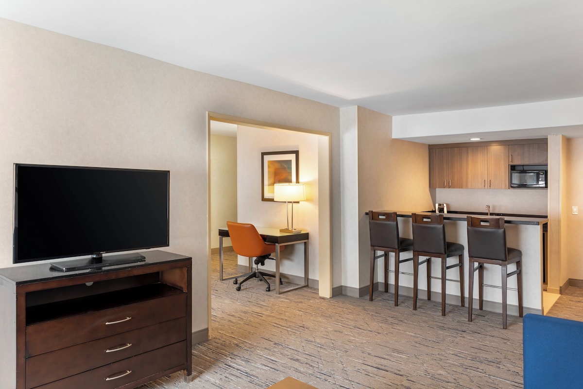 A hotel room with a flat-screen TV, a desk with a chair, a dining area with chairs, and a kitchenette with a microwave.