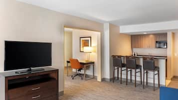 A hotel room with a flat-screen TV, a desk with a chair, a dining area with chairs, and a kitchen with a microwave.