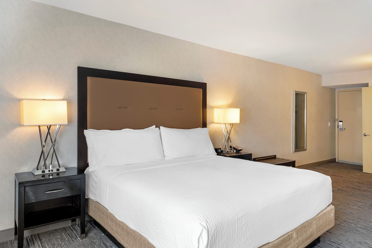 A hotel room with a large bed, two bedside tables with lamps, a nightstand, a mirror, and a door.