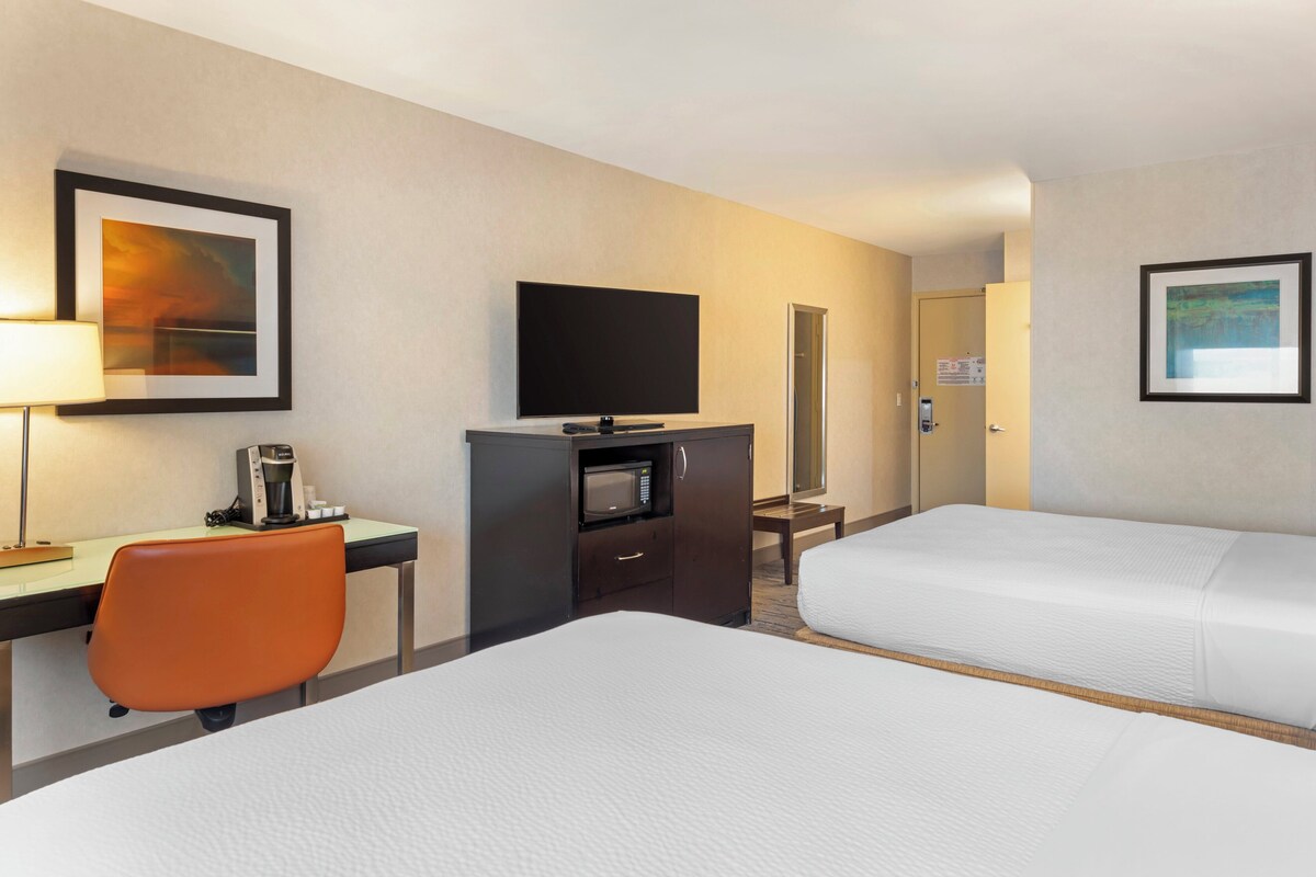 A hotel room with two beds, a desk with a coffee maker, a TV, and framed artwork on the walls.