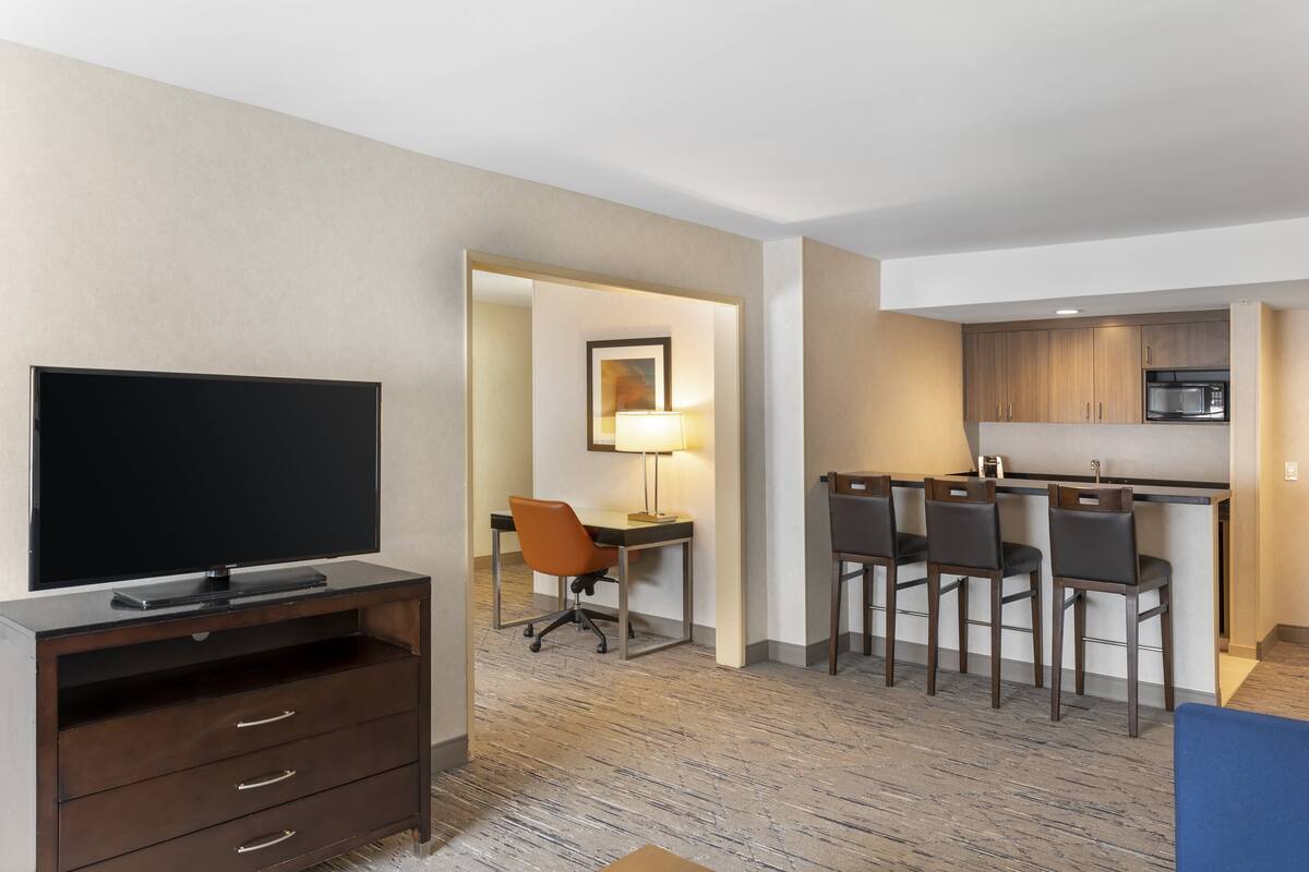 A hotel room with a flat-screen TV, a desk with a chair, a dining area with chairs, and a kitchenette with a microwave.