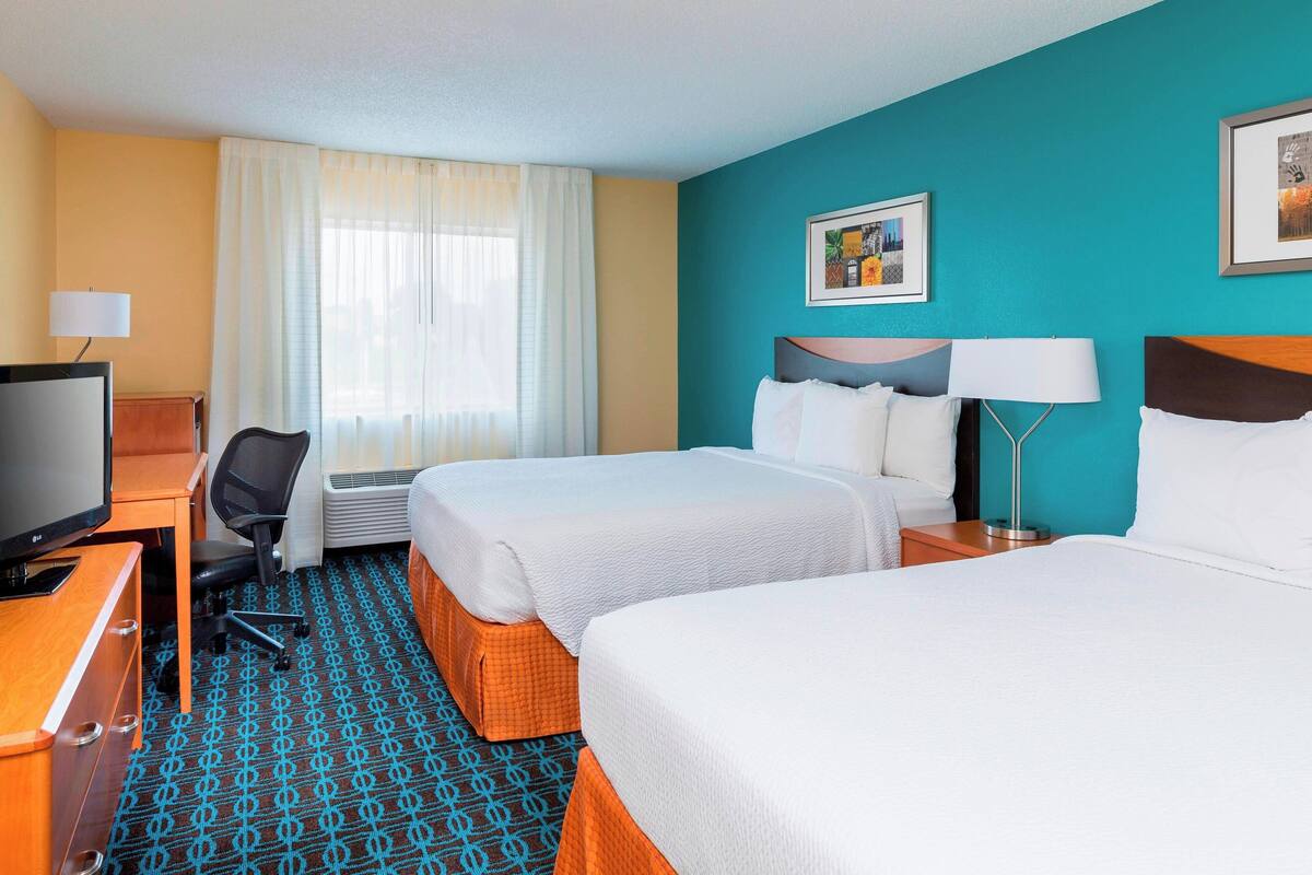 A hotel room with two beds, a desk with a television, a chair, and a window with curtains.