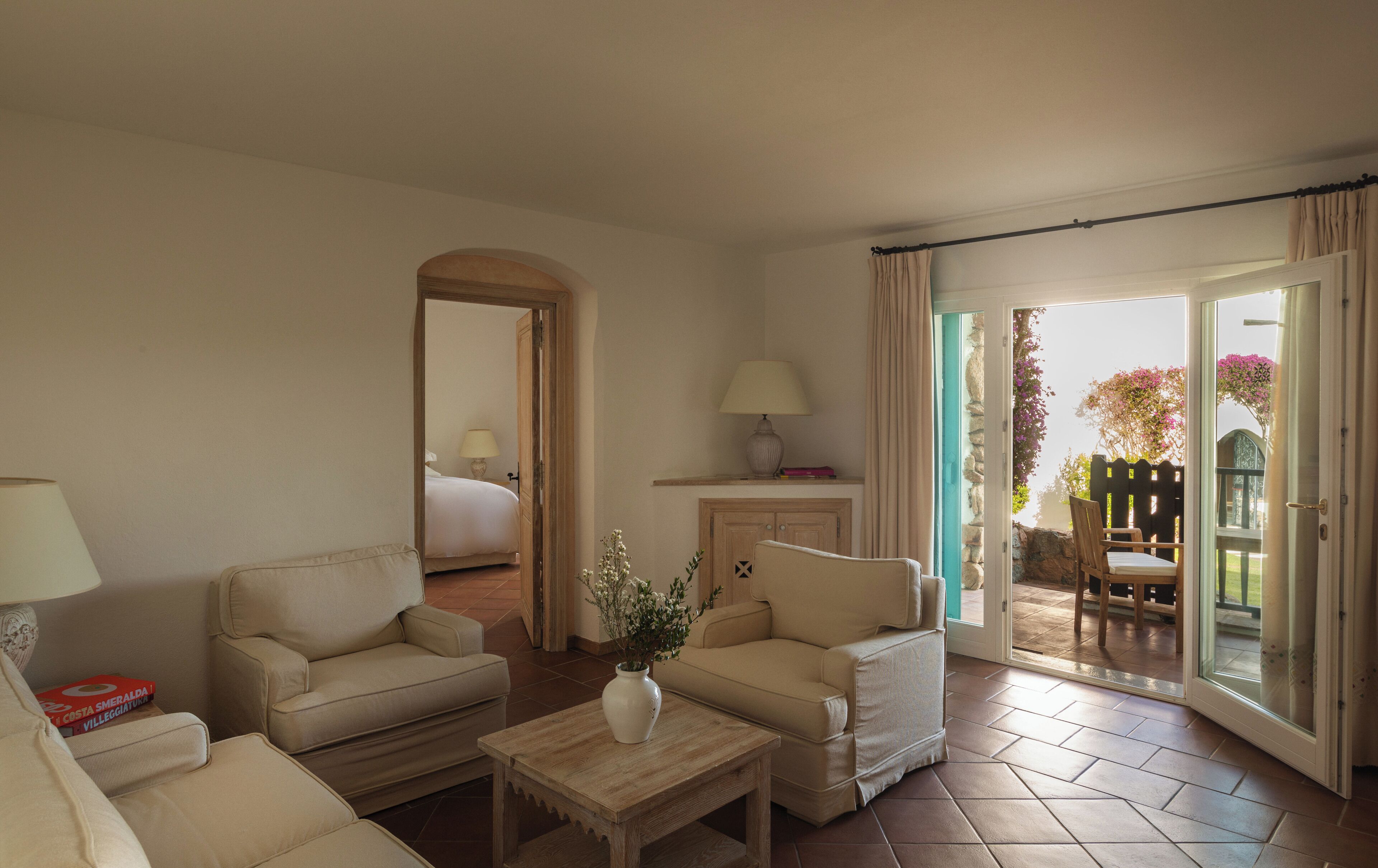 Ein Wohnzimmer mit einer Couch, einem Sessel und einem kleinen Tisch mit einer Vase. Eine Tür führt zu einem Balkon mit Blick auf einen Garten.