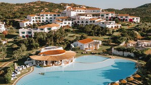 A large swimming pool with a circular deck, surrounded by lounge chairs and umbrellas, and a resort with multiple buildings and a mountainous backdrop. - Romazzino, A Belmond Hotel, Costa Smeralda (Arzachena)