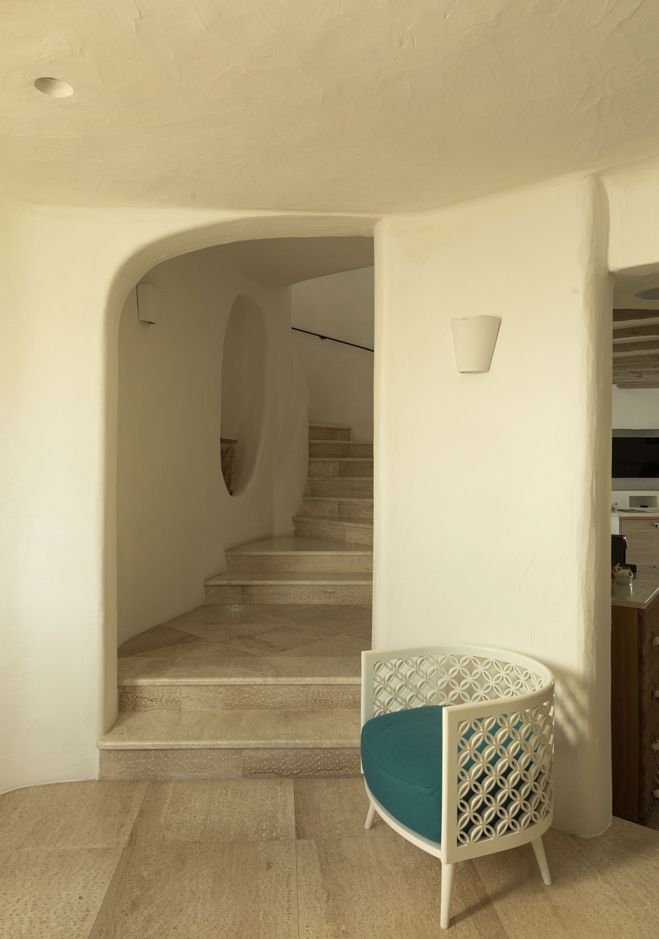 A modern interior with a staircase leading to an upper level, a white chair with a geometric pattern, and a light fixture on the wall.