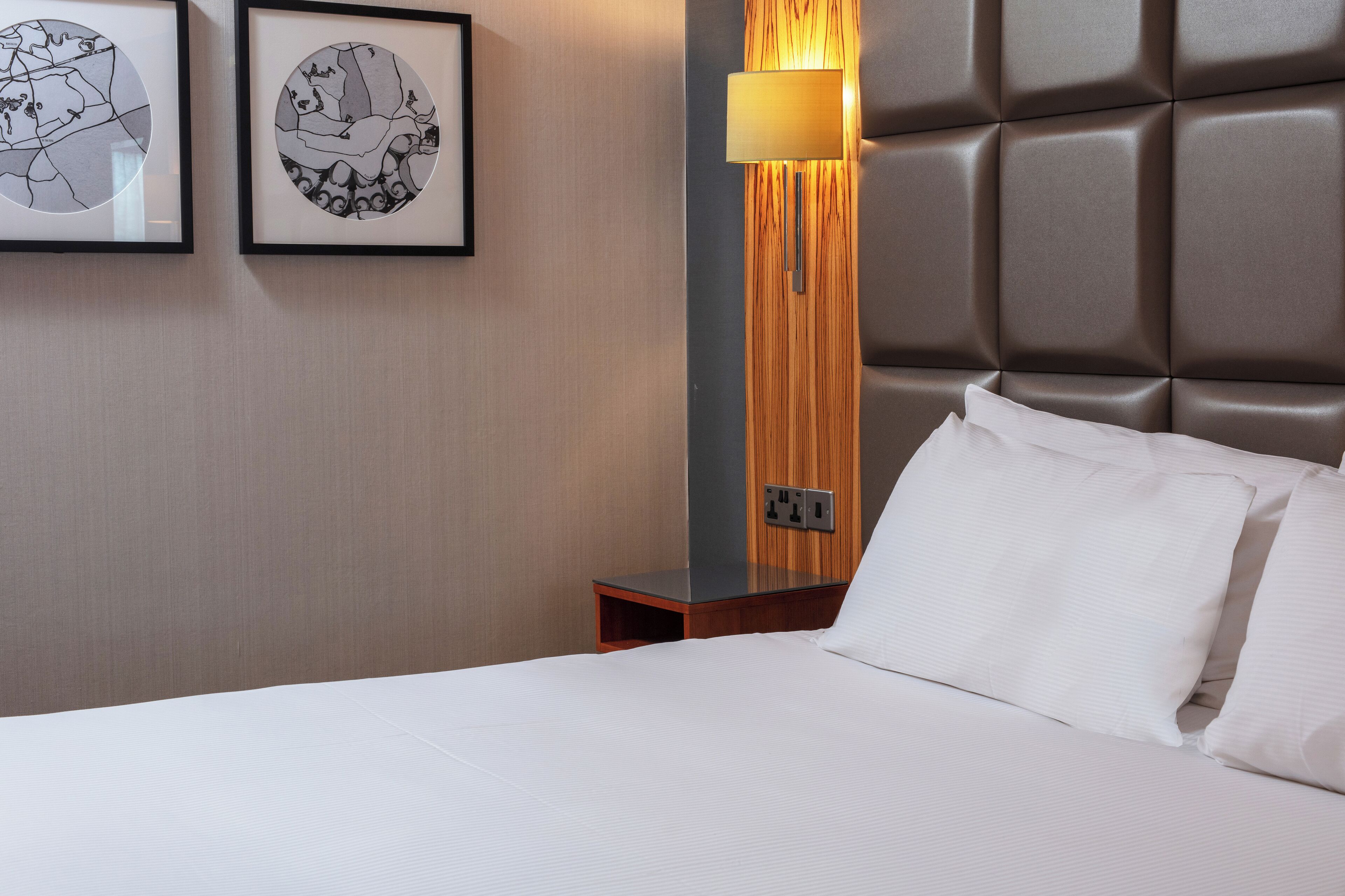 A hotel room with a large bed, a nightstand with a lamp, and two framed artworks on the wall.