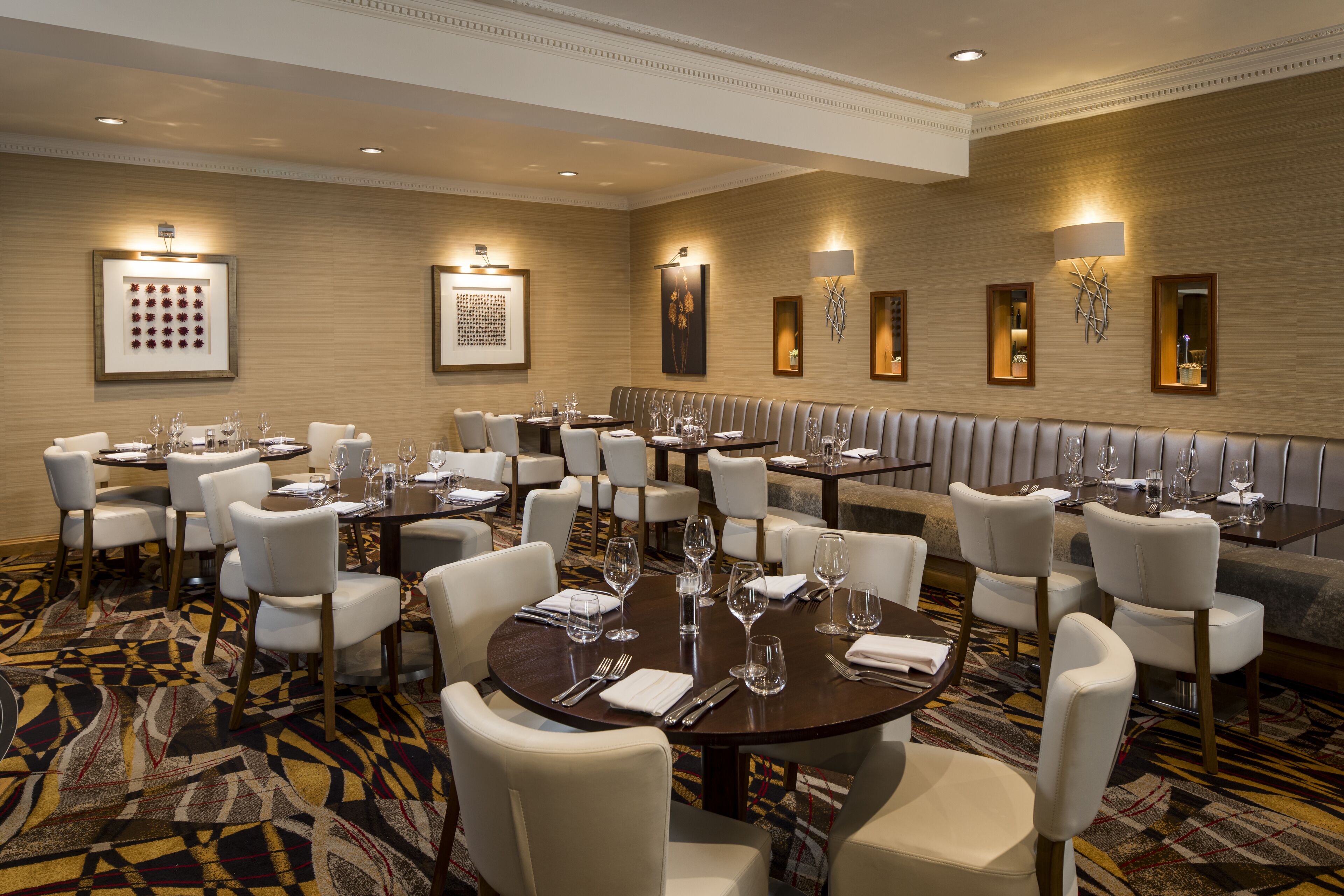 A hotel dining area with round tables and white chairs, framed artwork on the walls, and a patterned carpet.