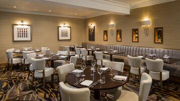 A hotel dining area with round tables and white chairs, framed artwork on the walls, and a patterned carpet.