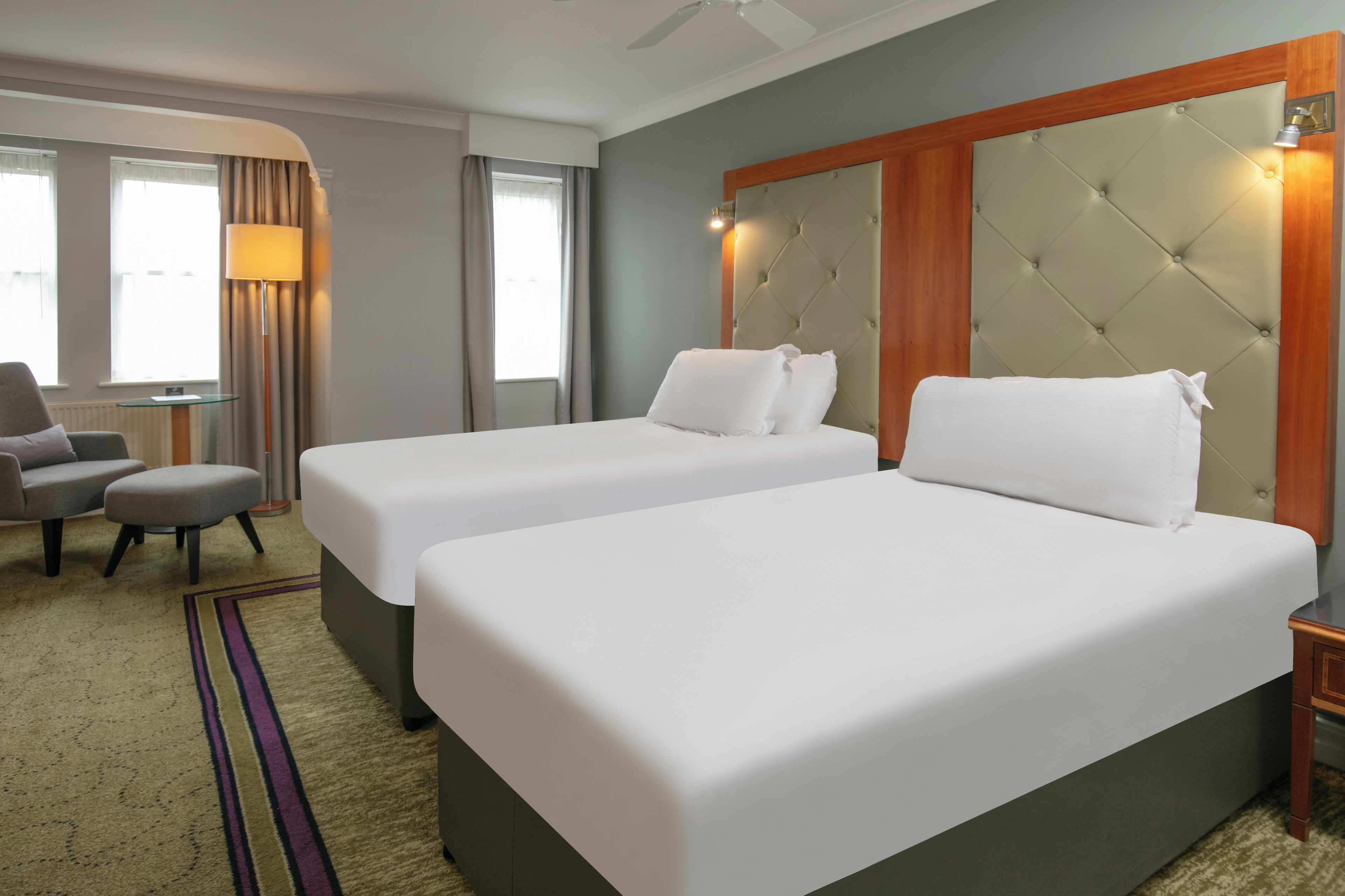 A hotel room with two single beds, a desk, a chair, and a lamp.