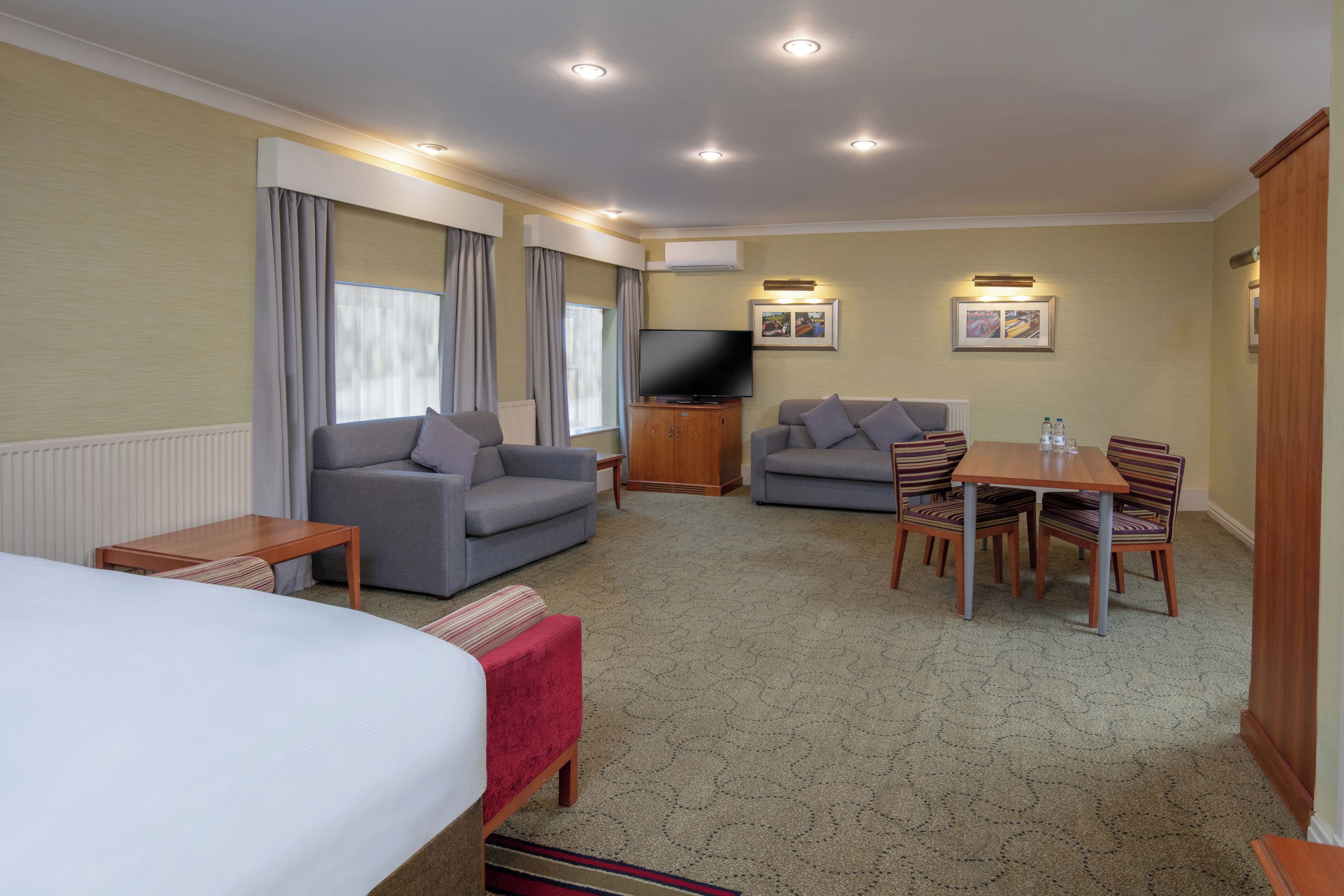 Habitación de hotel con cama, dos sofás, televisor, mesa de comedor con sillas y piso alfombrado.