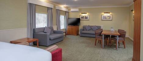 A hotel room with a bed, two sofas, a TV, a dining table with chairs, and a carpeted floor.