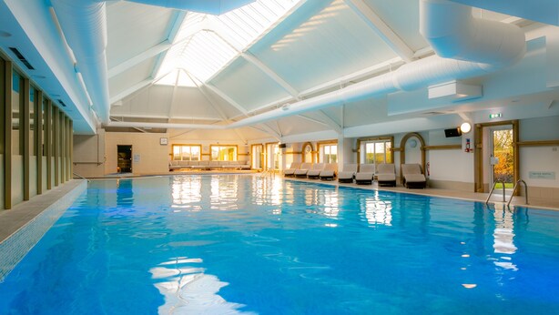 An indoor swimming pool with clear blue water, a high ceiling with skylights, and a row of lounge chairs along the side. - The Park Royal Hotel & Spa (Warrington)