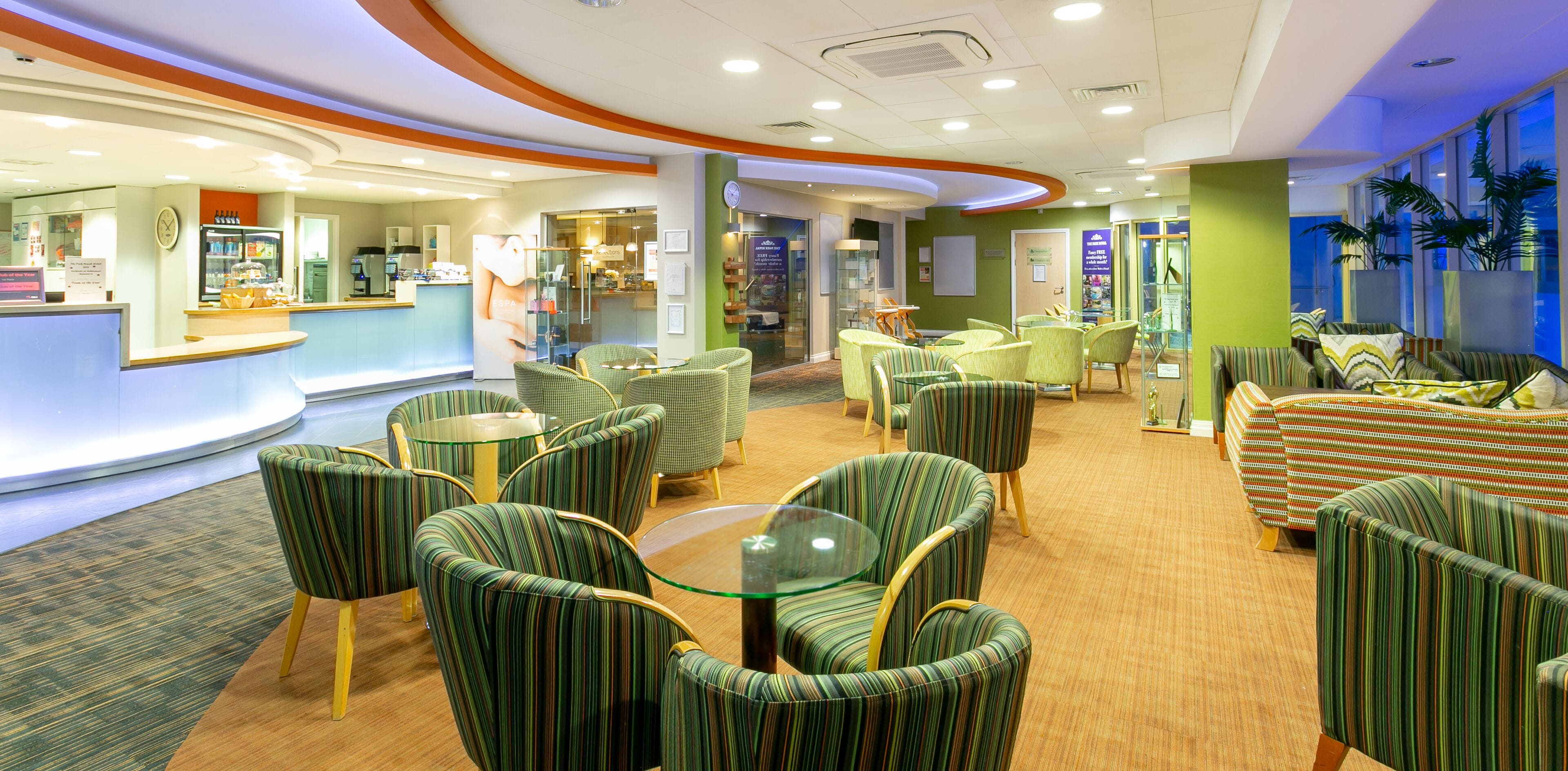 A hotel lobby with a reception desk, seating area with round tables and chairs, and a green wall with a clock.