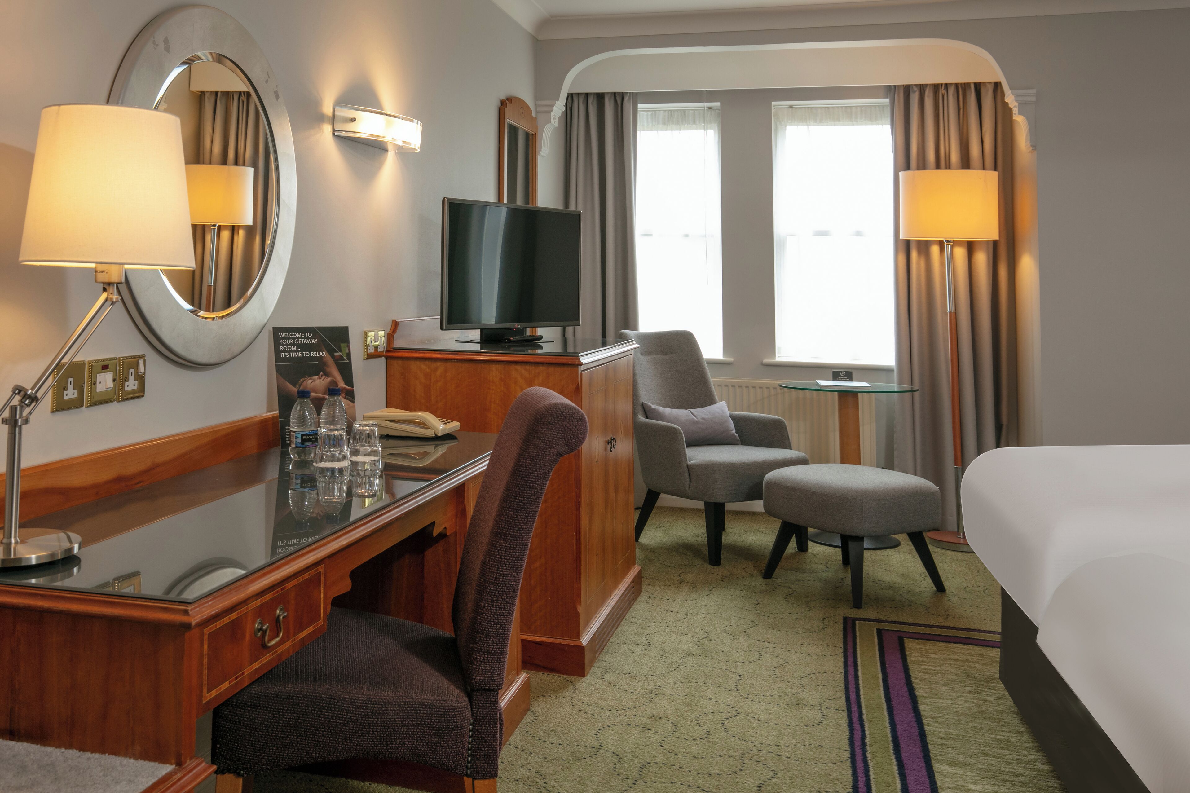 Una habitación de hotel con cama, escritorio con computadora, televisor, silla y pequeña mesa con lámpara.