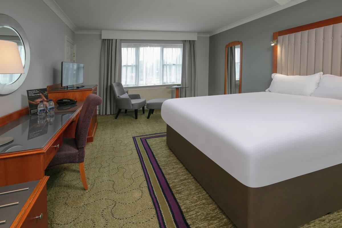 A hotel room with a large bed, a desk with a computer, a chair, and a window with curtains.