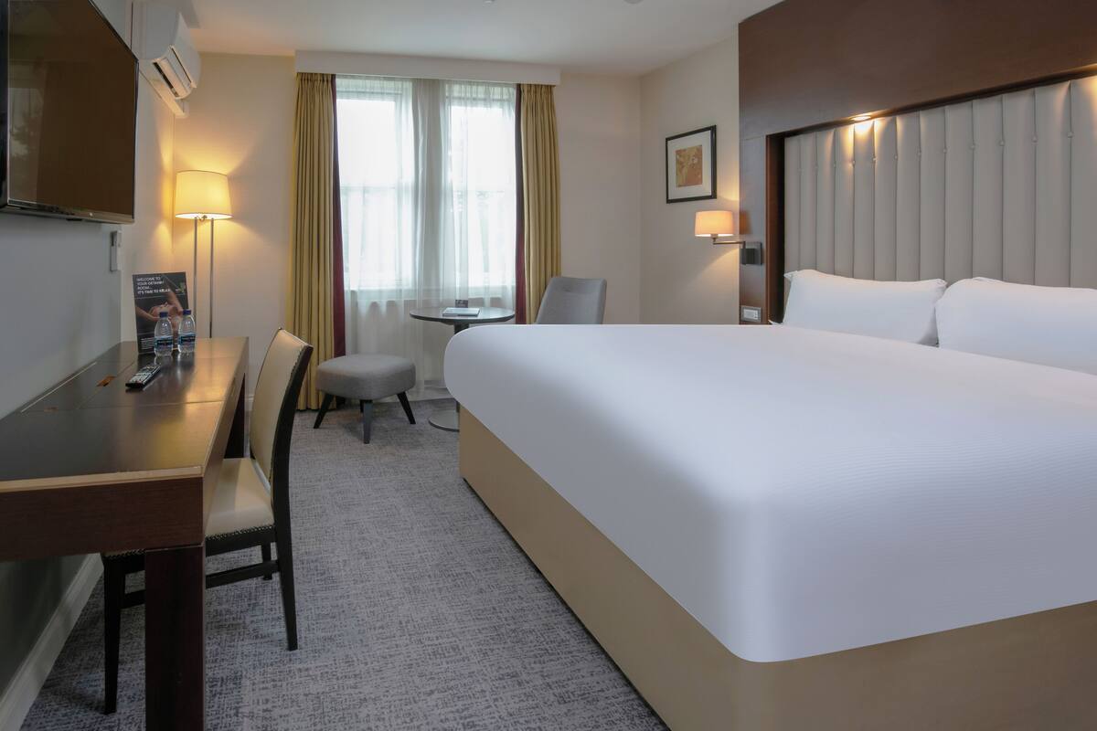 A hotel room with a large bed, a desk with a chair, a small table, and a TV.