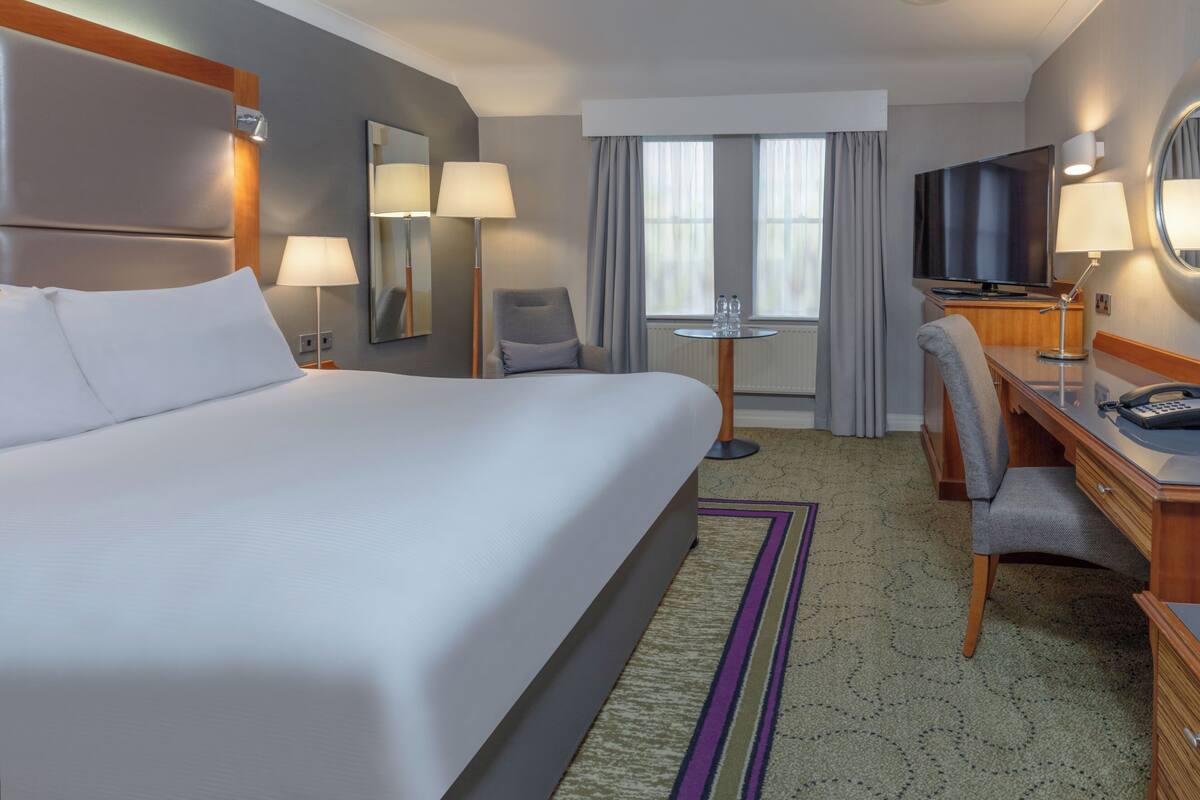 A hotel room with a large bed, a desk, a chair, a television, and a small table with two lamps.