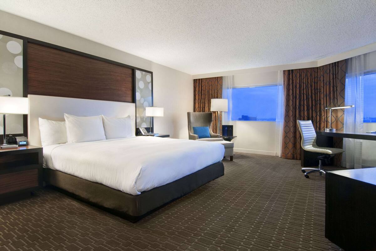 Una habitación de hotel con una cama grande, un escritorio, una silla y un ventanal con cortinas.