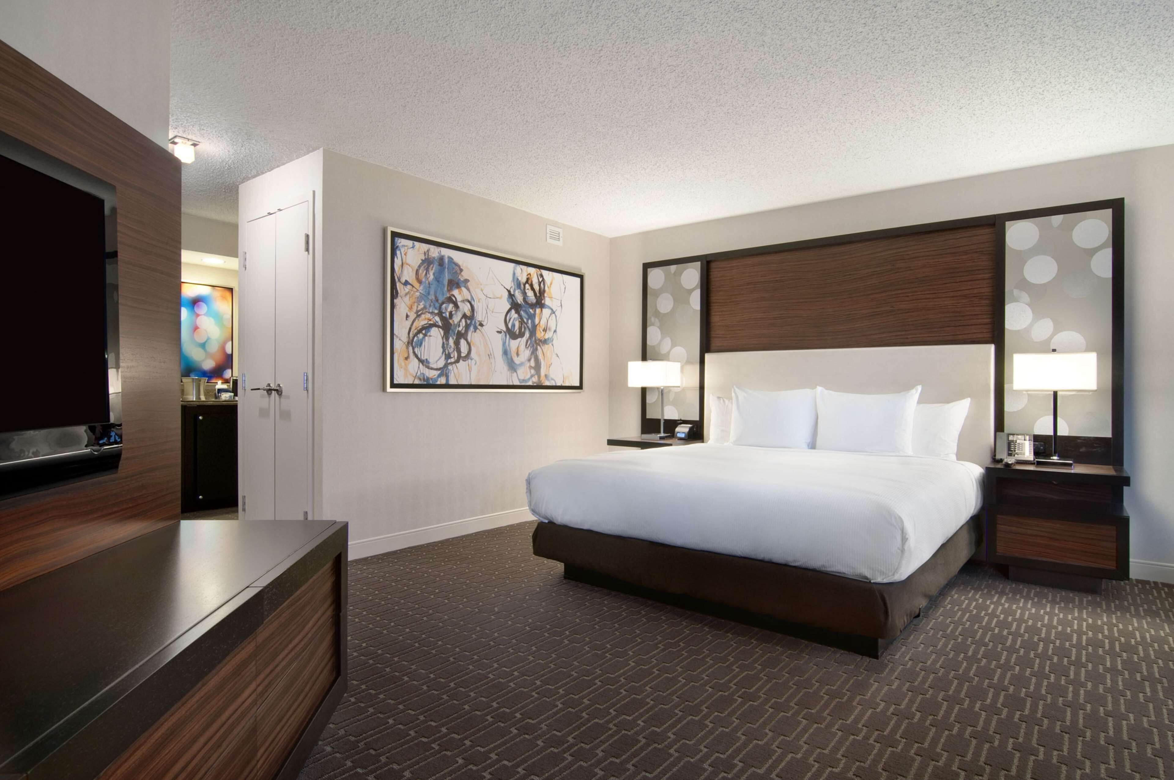 A hotel room with a large bed, a TV, artwork on the wall, and a patterned carpet.