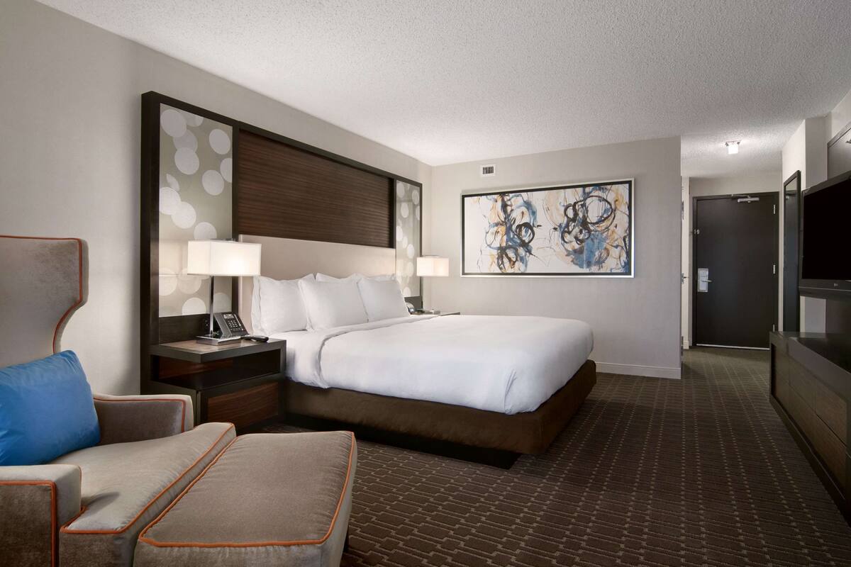 Habitación de hotel con una cama, un escritorio con lámpara, una silla, un televisor y un cuadro abstracto grande en la pared.