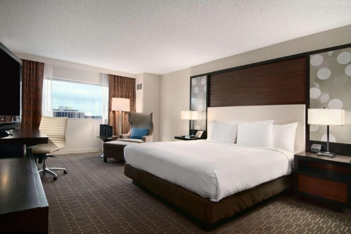 A hotel room with a large bed, a desk, a chair, two bedside tables with lamps, and a view of the city through a window.