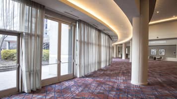 A spacious corridor with large windows, a patterned carpet, and a grand salon in the distance.