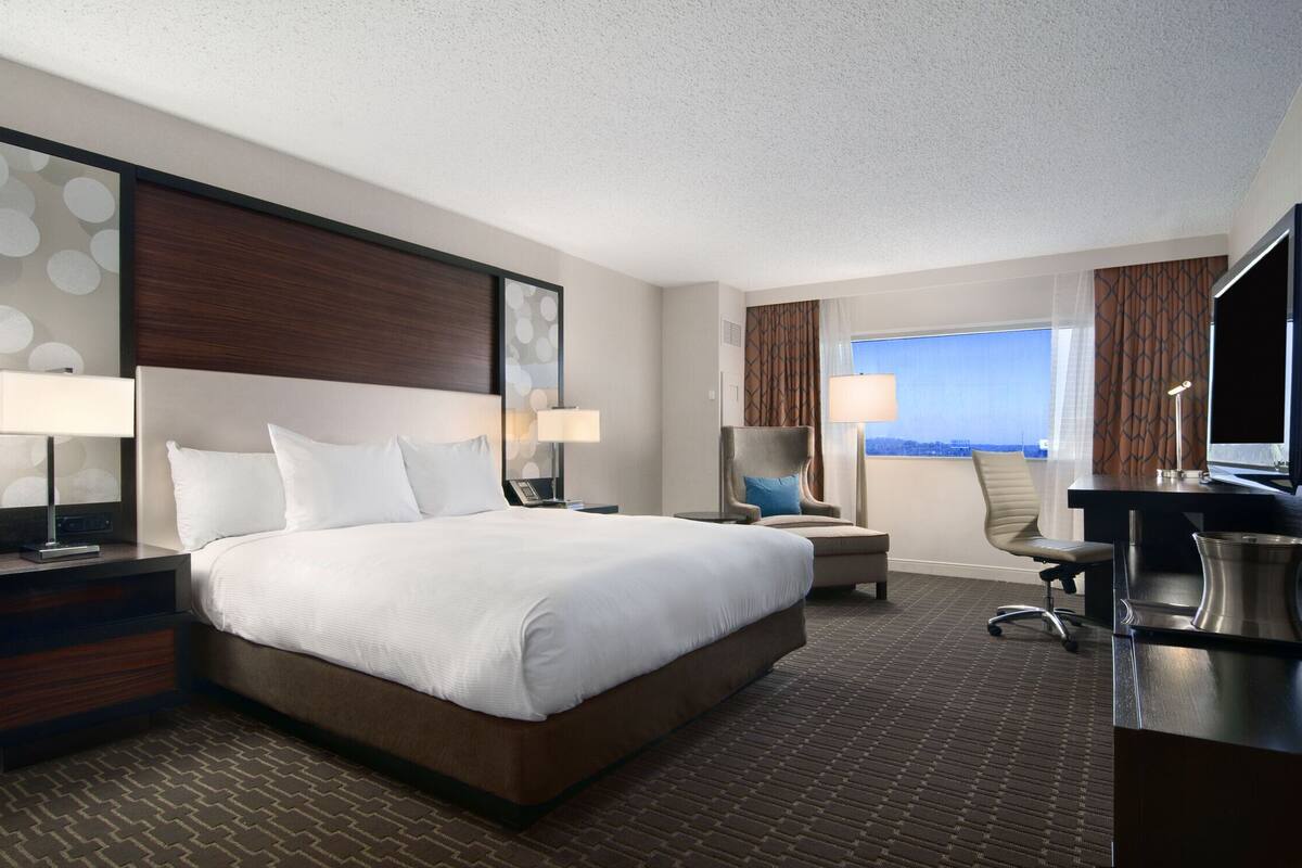 Habitación de hotel con una cama grande, un escritorio con computadora, una silla y vista a la ciudad.