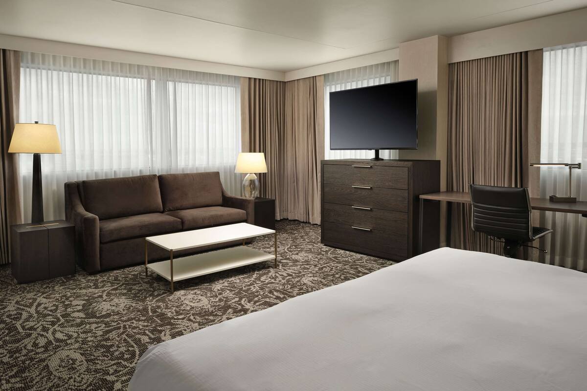 Habitación de hotel con una cama grande, un sofá, un escritorio con silla, un televisor de pantalla plana y dos lámparas.