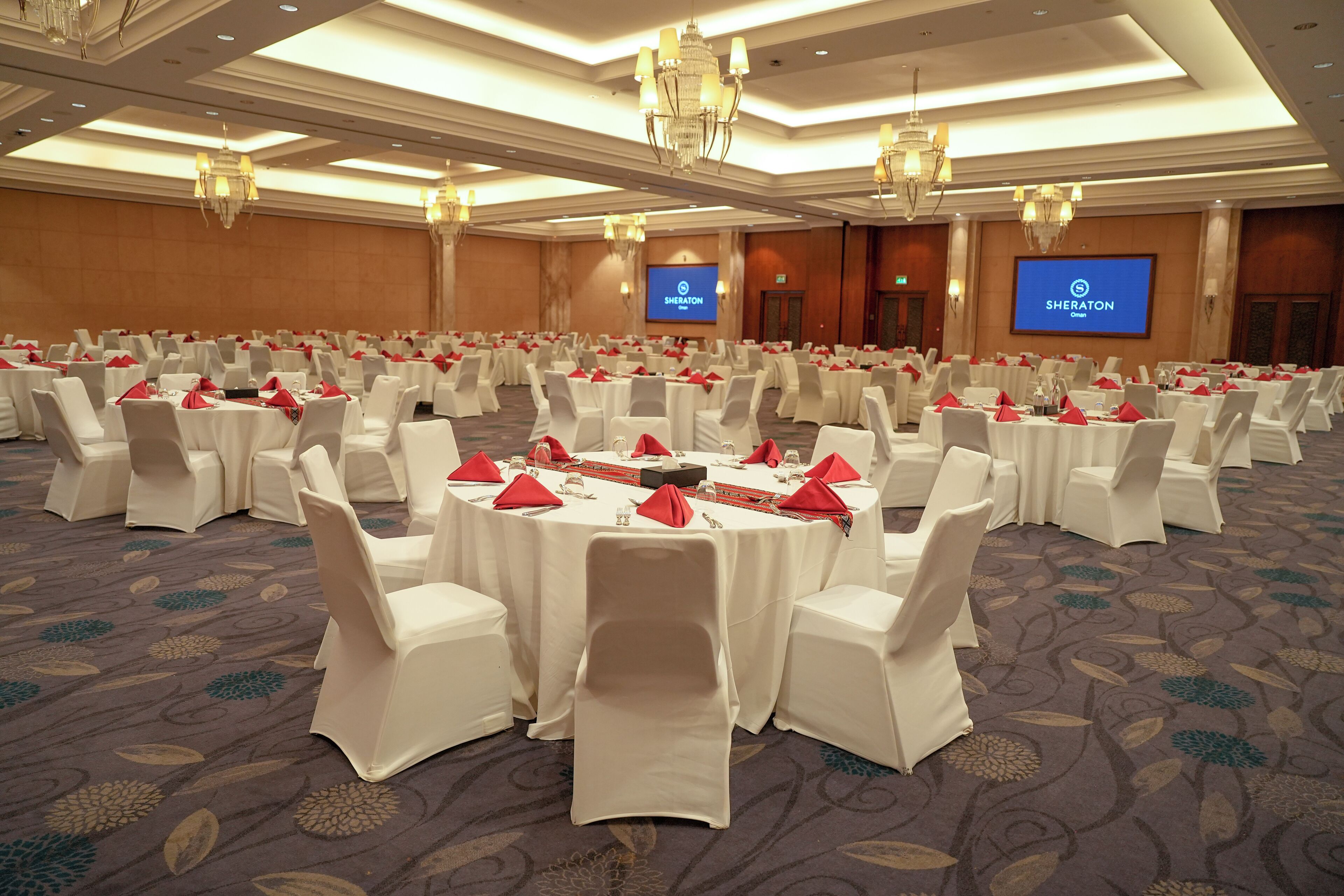Une grande salle de conférence avec des tables rondes, des chaises blanches et des serviettes rouges.