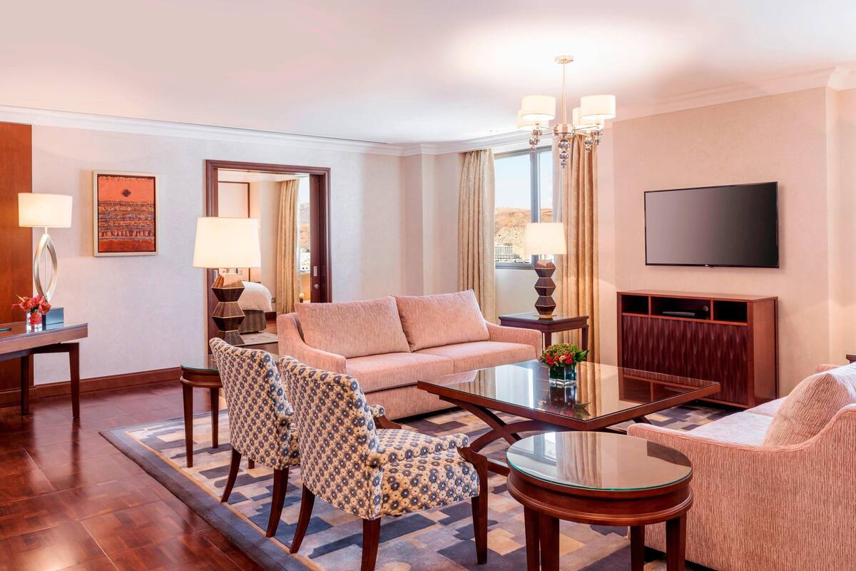 Habitación de hotel con sofá, dos sillones estampados, una mesita de centro, televisor y un espejo grande.