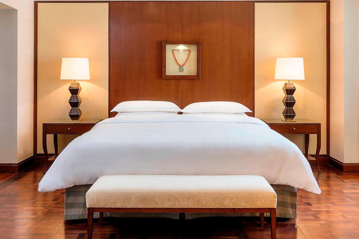 Una cama grande con ropa de cama blanca, dos mesitas de noche con lámparas, un banco al pie de la cama y un cuadro en la pared.
