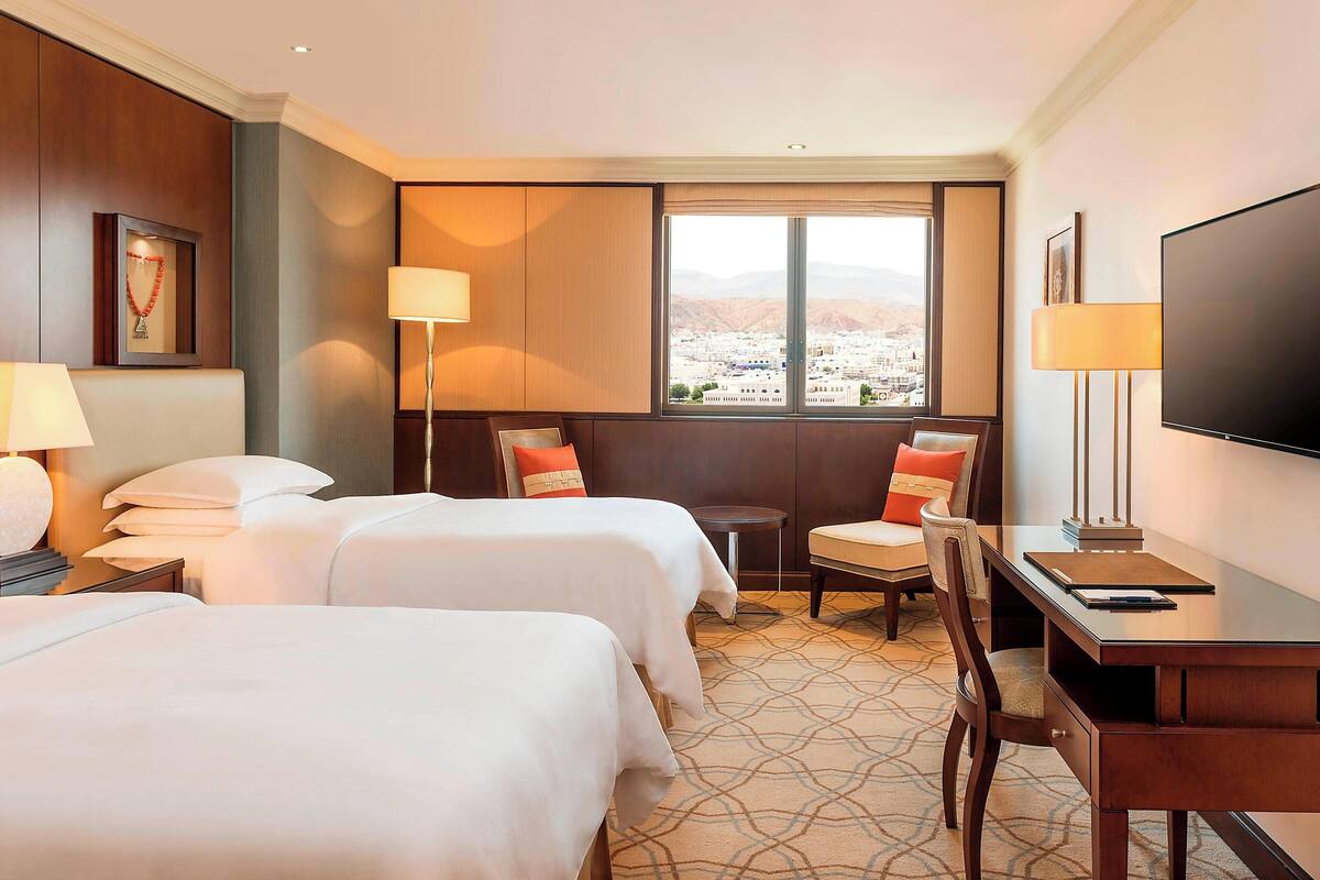 Habitación de hotel con dos camas, un escritorio, una silla, un televisor y una ventana con vistas a la ciudad.