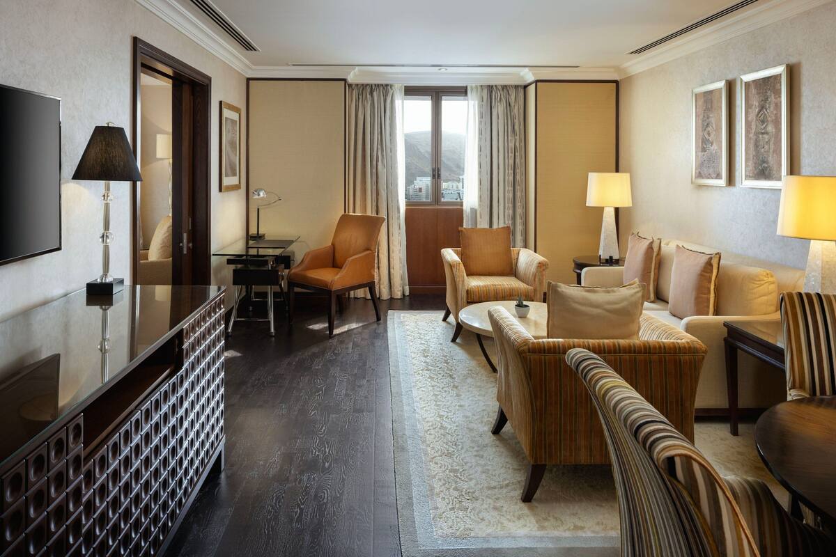 Habitación de hotel con televisión de pantalla plana, una mesa de centro de vidrio, un sofá, una silla y una pequeña mesa con lámpara.