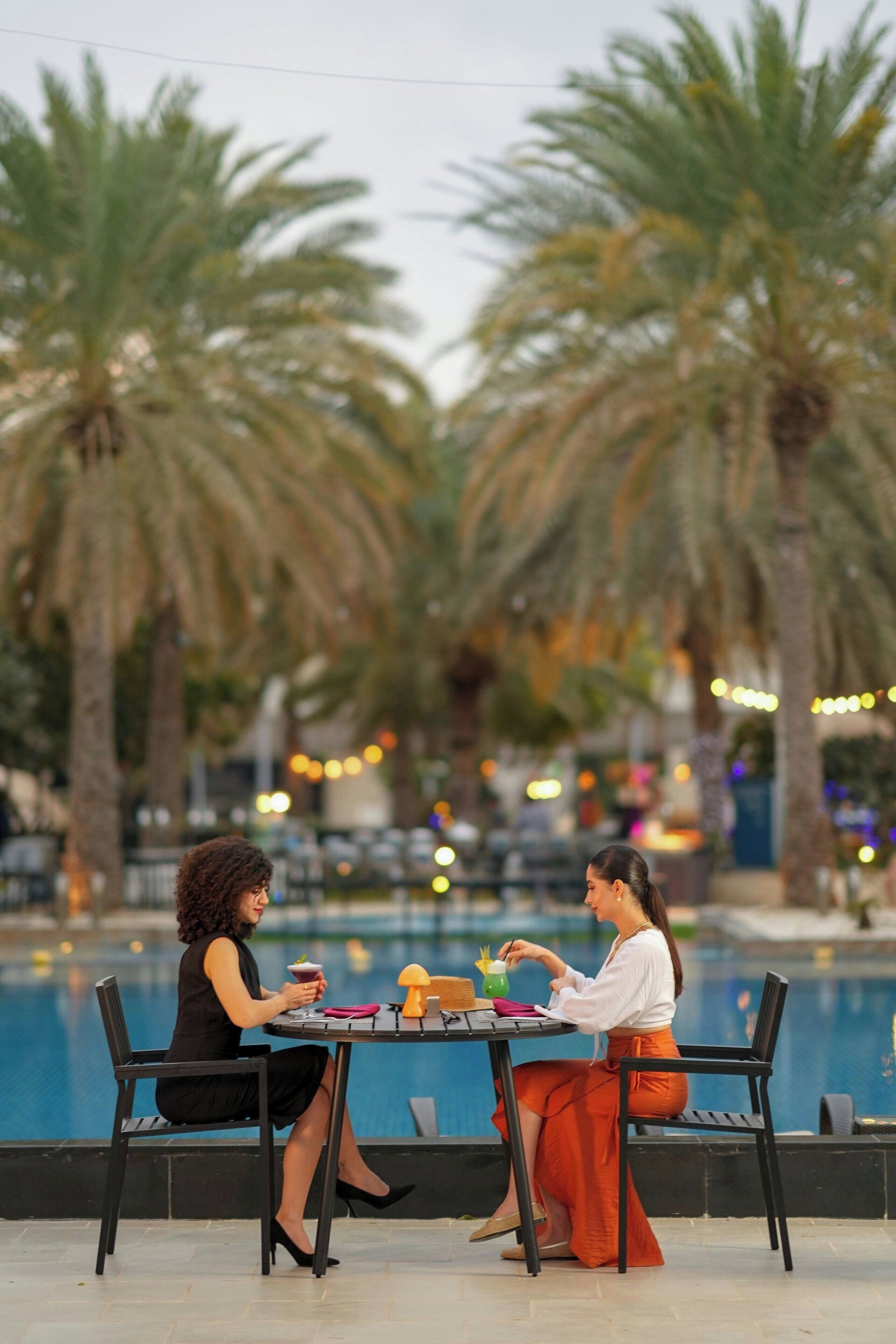 Two people seated at a table by a poolside with palm trees in the background.