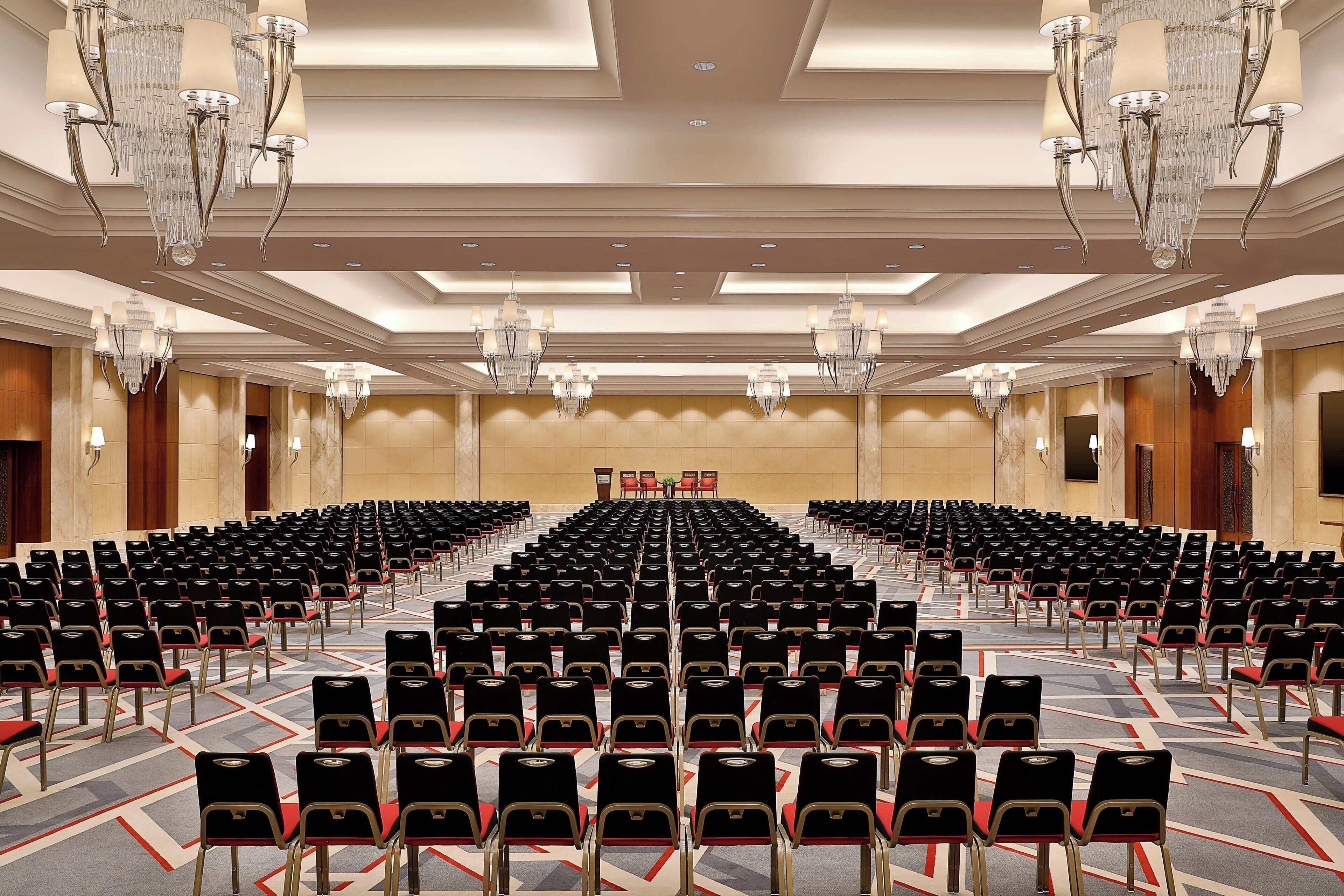Een ruime congreszaal met rijen zwarte stoelen, een podium aan de voorzijde en kroonluchters.