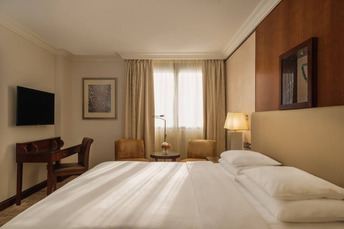 Habitación de hotel con una cama grande, un escritorio, dos sillas, un televisor, una lámpara y un cuadro en la pared.