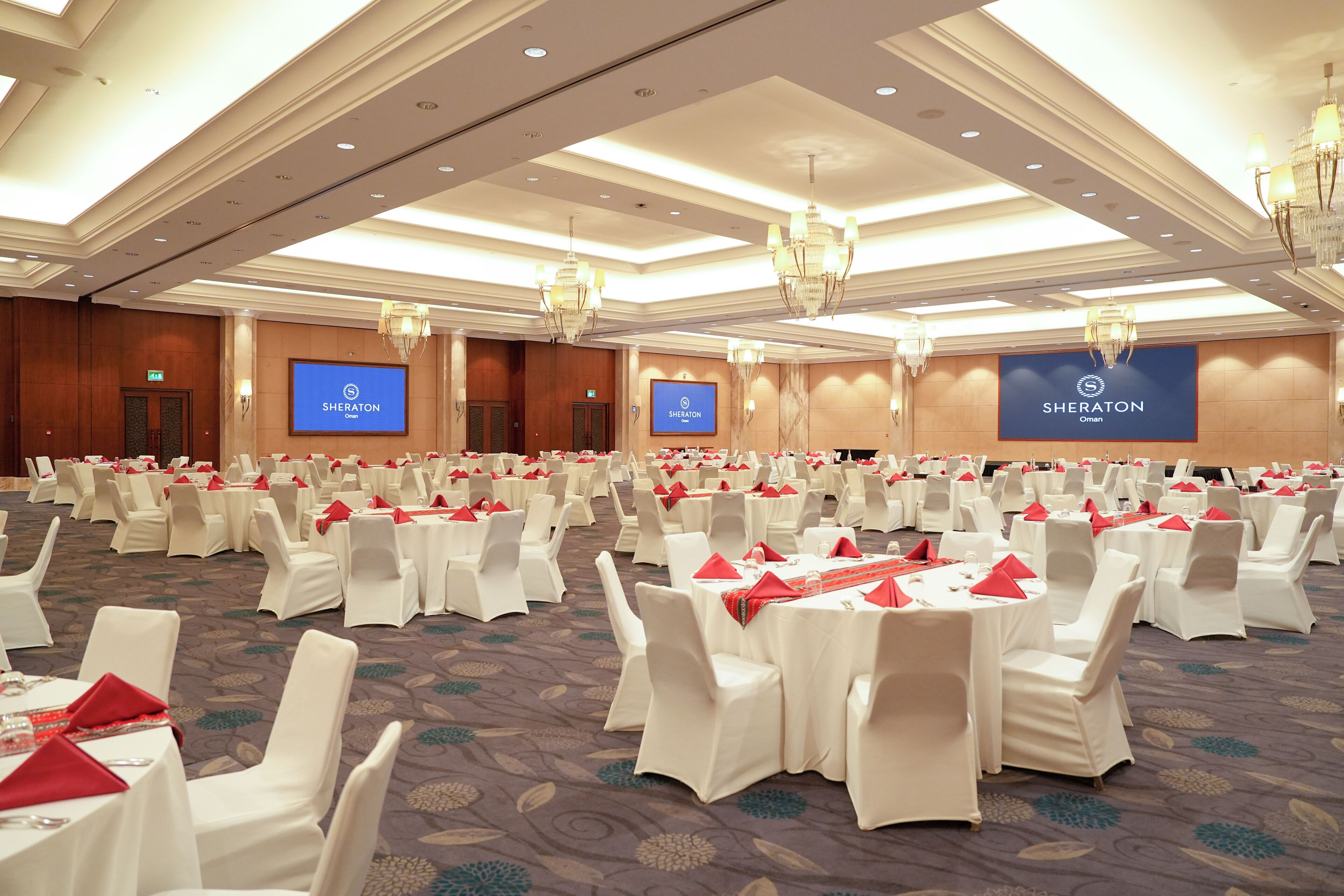A Sheraton hotel ballroom with multiple round tables and white chairs, each with red napkins.