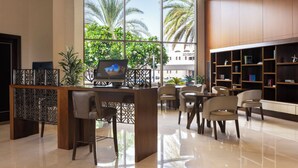 A modern hotel lobby with a reception desk, seating area, and a large window offering a view of palm trees.